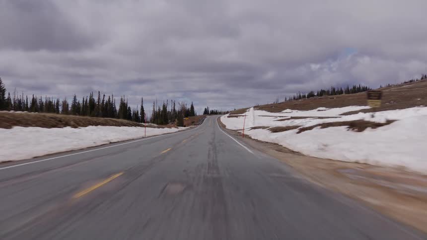 A point-of-view shot driving along a winding mountain highway with melting snow on the sides under a cloudy sky