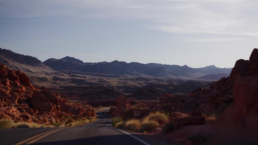 A slow, smooth shot travels along a winding desert road, revealing a vast valley of red rock formations and mountains at sunrise