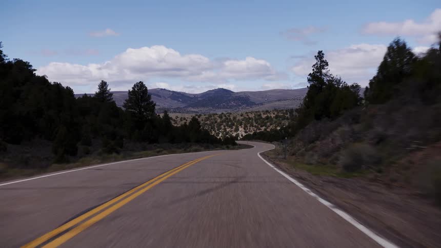 POV shot driving along a winding two-lane road that meanders through a vast and scenic mountain landscape under a partly cloudy sky