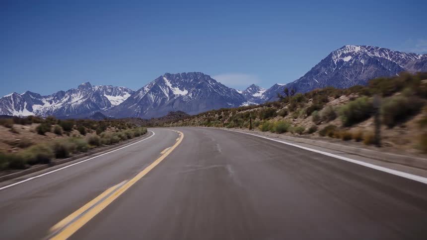 Driving POV along a winding desert highway towards the majestic snow-capped Sierra Nevada mountains on a clear day