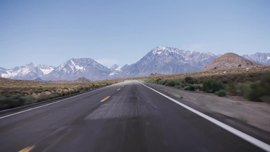 Driving down a desolate desert highway towards a majestic range of snow-capped mountains on a clear day