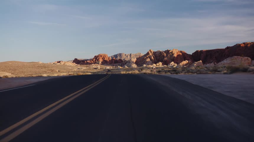 A point-of-view shot driving over a hill on a desert road, revealing a scenic landscape of colorful rock formations at sunrise