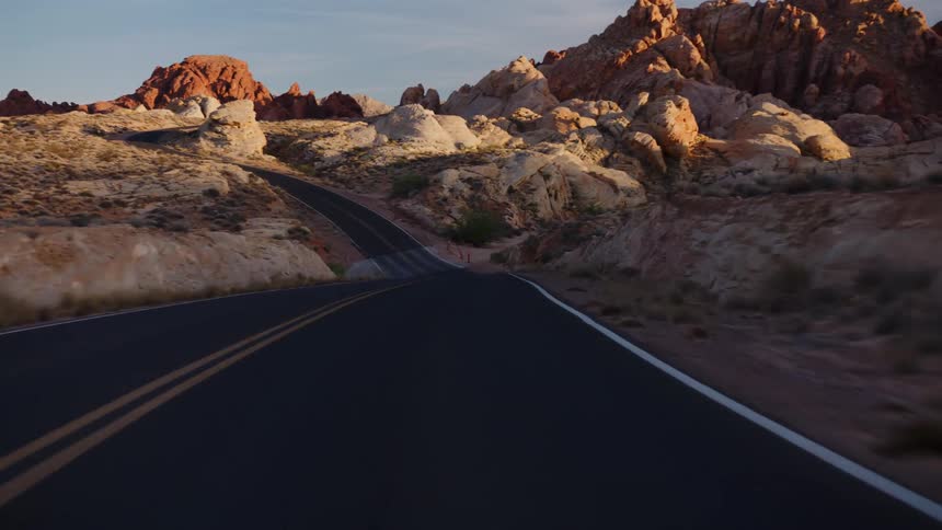 A point-of-view drive on a winding road through a desert canyon with red rock formations at sunset