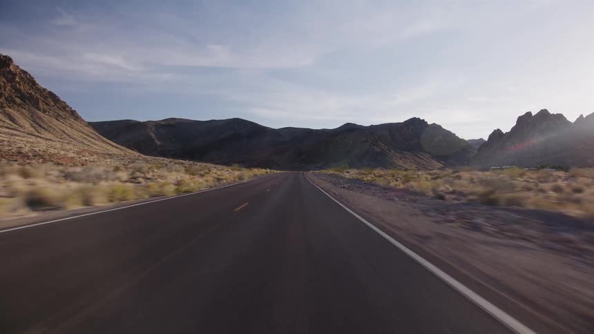 A point-of-view driving shot on a lonely desert highway surrounded by majestic mountains during a beautiful sunset