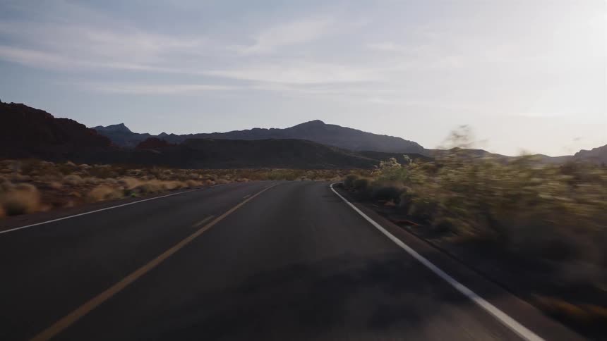 A point-of-view shot driving along a winding two-lane highway through a vast desert landscape with mountains in the distance at sunrise
