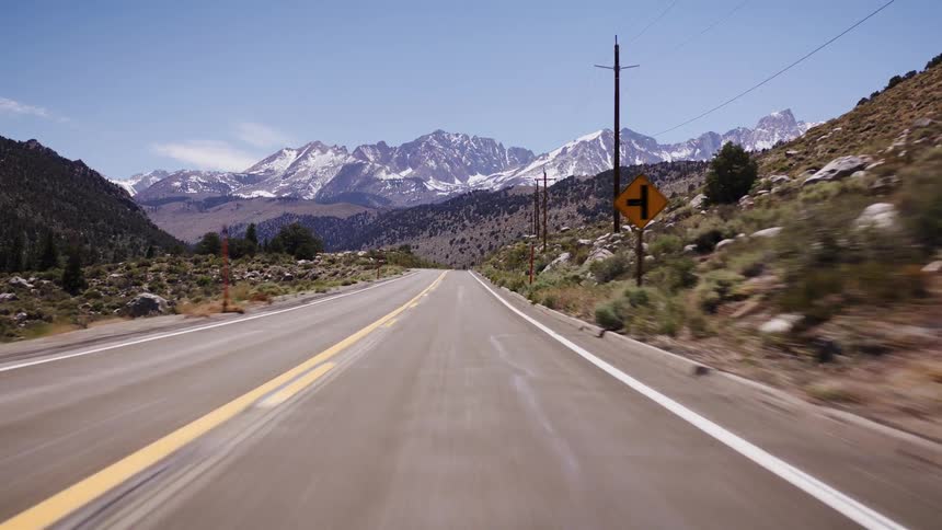 A POV driving shot on a scenic mountain highway heads toward a snow-capped mountain range on a sunny day