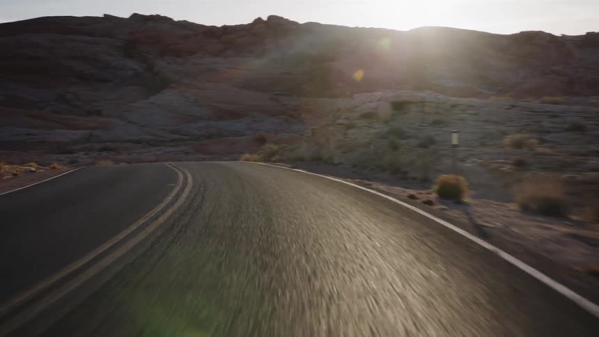 A point-of-view perspective driving down a winding asphalt road through a vast desert landscape with unique red rock formations at golden hour