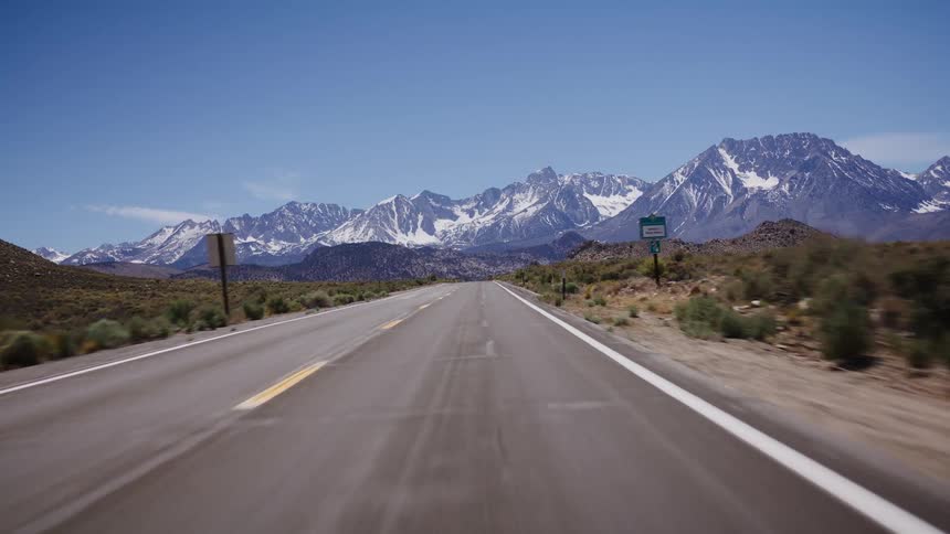 A smooth point-of-view drive down a long, straight desert highway leading toward a beautiful snow-capped mountain range under a clear blue sky