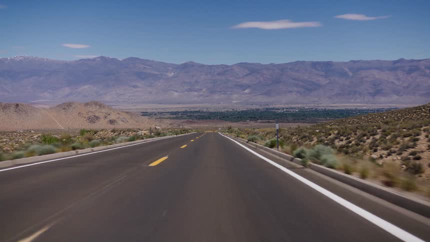 A point-of-view perspective driving down a long, straight desert highway towards a distant mountain range and valley