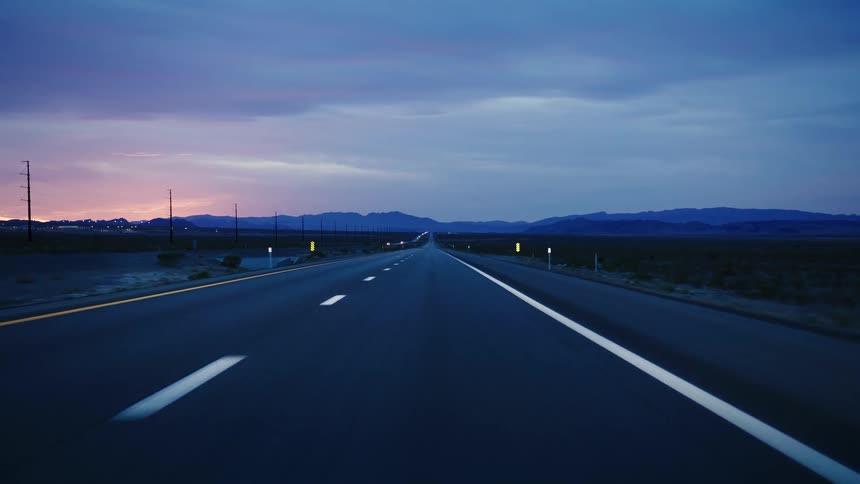 A point-of-view shot driving down a long, empty highway through the desert towards distant mountains at twilight