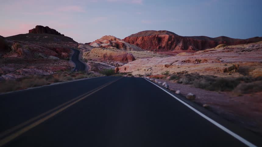 A slow, first-person drive along a winding desert highway surrounded by vibrant red and orange rock formations at dusk
