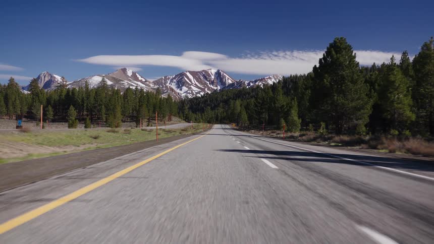 A first-person view of driving along a scenic highway towards majestic snow-capped mountains on a clear sunny day