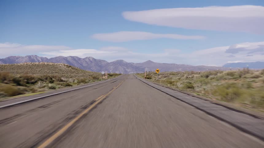 A driver's point-of-view shot traveling down a long, empty desert highway toward distant mountains before tilting up to the clear blue sky