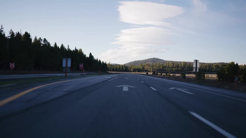 A serene point-of-view shot driving down an open highway surrounded by lush forests and rolling hills under a bright blue sky
