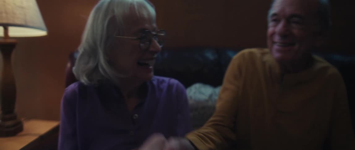 An elderly woman with glasses shakes hands and smiles warmly next to her husband in their cozy living room