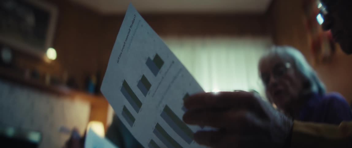 A senior man with glasses carefully reviews a printed document with bar charts, discussing the financial data with others in a dimly lit room