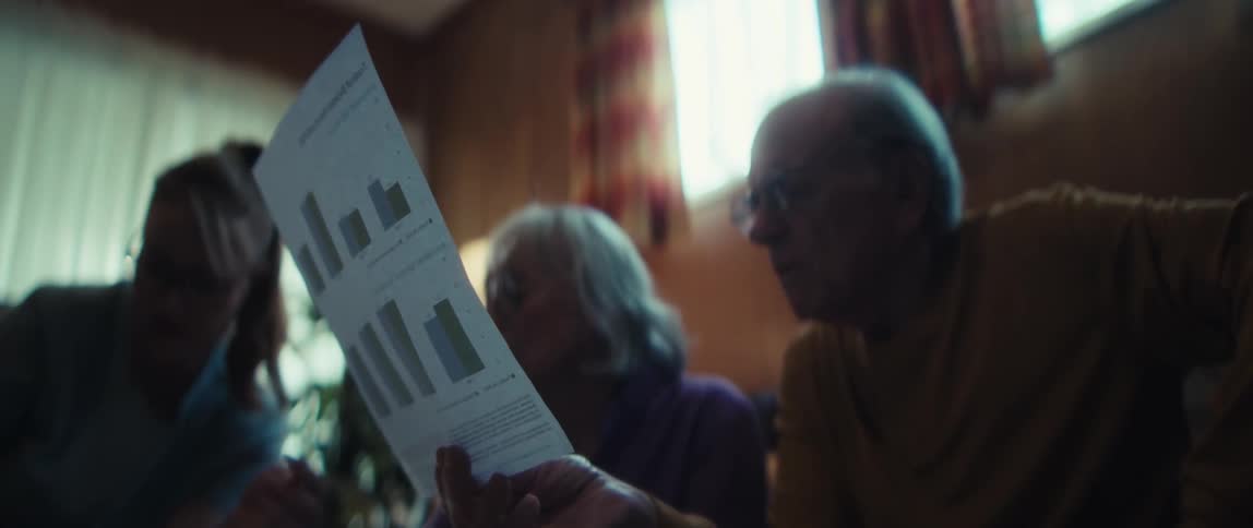 An elderly couple and a younger woman sit together in a dimly lit room, seriously reviewing a document with bar charts
