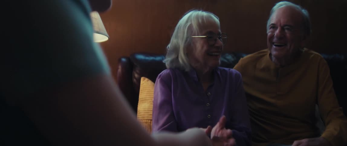 An elderly couple smiles and laughs while having a heartfelt conversation with a visitor in their cozy living room