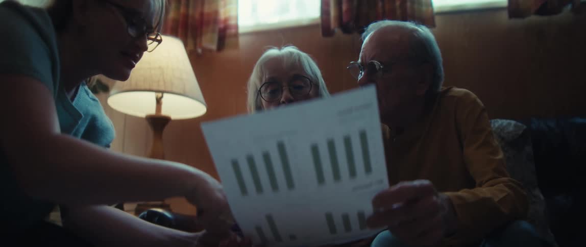 A caring woman explains a financial chart with bar graphs to a senior couple sitting on a couch in their living room