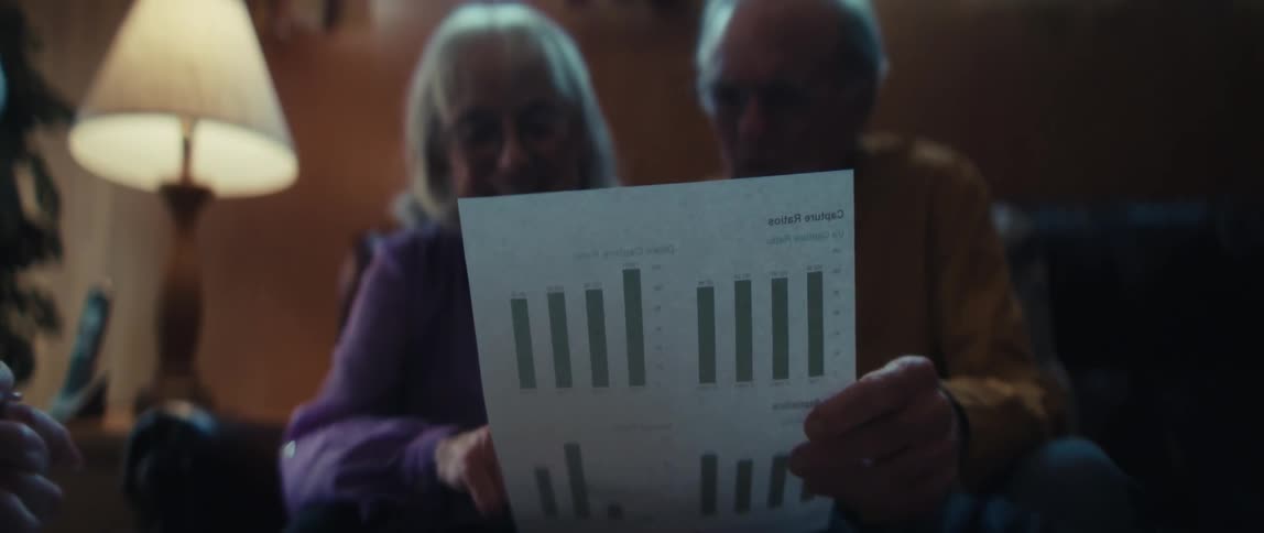 An elderly couple sits together at home, their initial smiles fading to looks of worry and concern as they review a financial document with bar charts