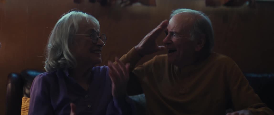 A senior couple greets a visitor with a handshake before bursting into joyful laughter while listening to a story