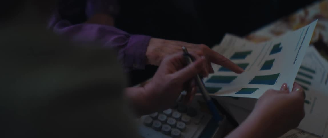 An older woman's hand points to a bar chart as a younger woman with a pen reviews the financial data together with a calculator