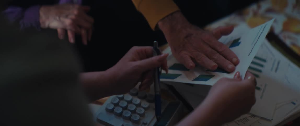 An older man's hand points to a bar chart while discussing financial data with a woman