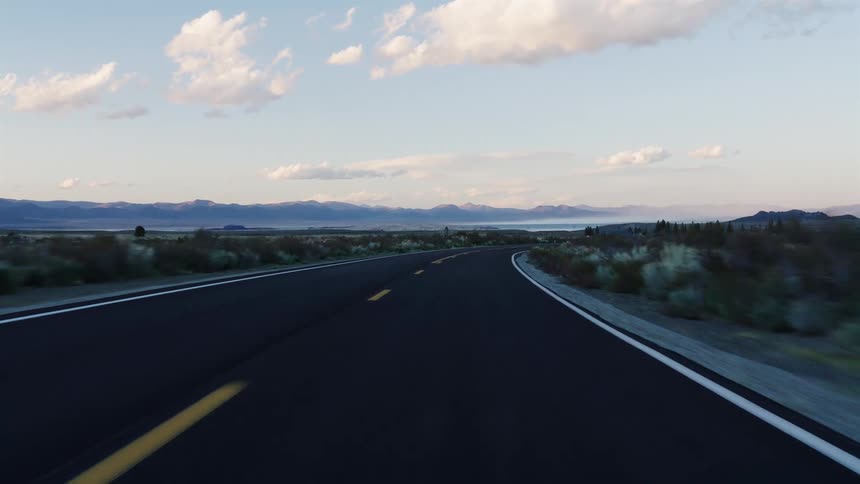 A point-of-view shot driving down a desolate desert highway towards distant mountains at dusk