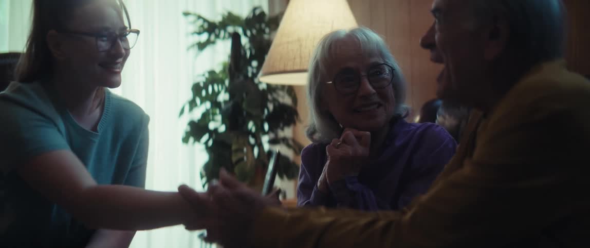 A young woman shares exciting news from a document with an elderly couple, resulting in joyful celebration, handshakes, and hugs in their home