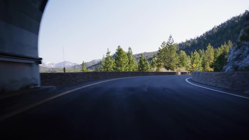 A point of view shot driving out of a dark tunnel and onto a scenic, winding mountain road on a sunny day