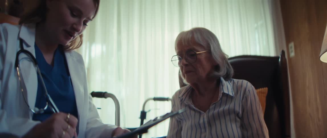 A friendly doctor consults with an elderly female patient during a home visit, taking notes on a clipboard