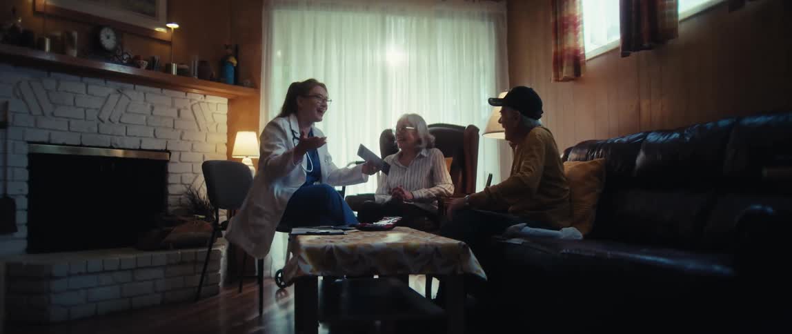 A female doctor on a house call laughs and talks with an elderly couple in their living room