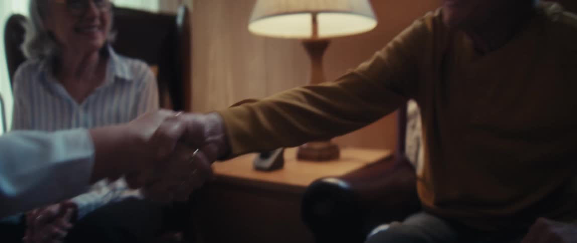 A home care provider warmly shakes the hand of a smiling elderly Vietnam veteran as his wife looks on
