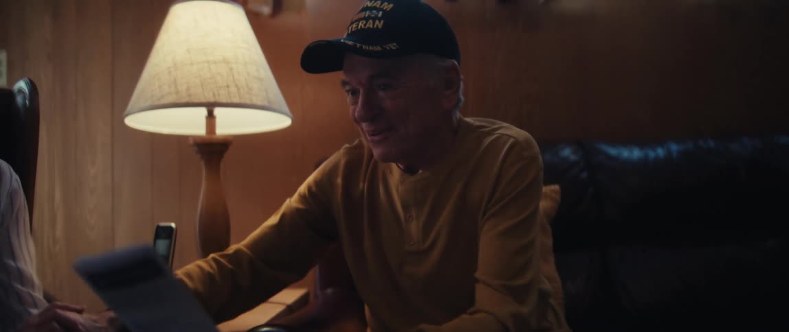 A grateful elderly veteran wearing a military cap shakes hands with a visitor in his home, with his smiling wife beside him
