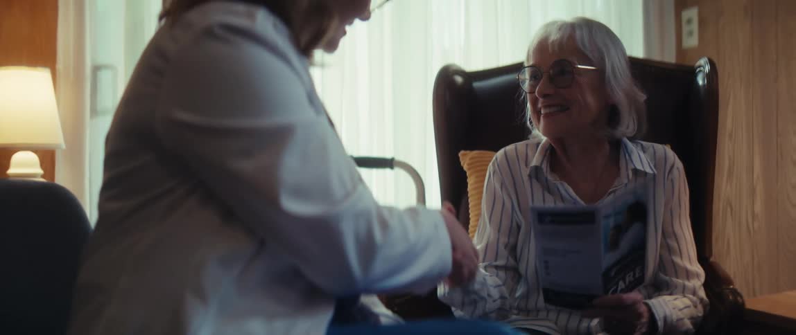 A smiling senior woman holds a care brochure and shakes hands with a healthcare worker during a home consultation
