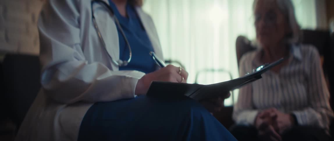 A dedicated female doctor in a white coat takes notes on a clipboard while consulting with an elderly patient during a home visit