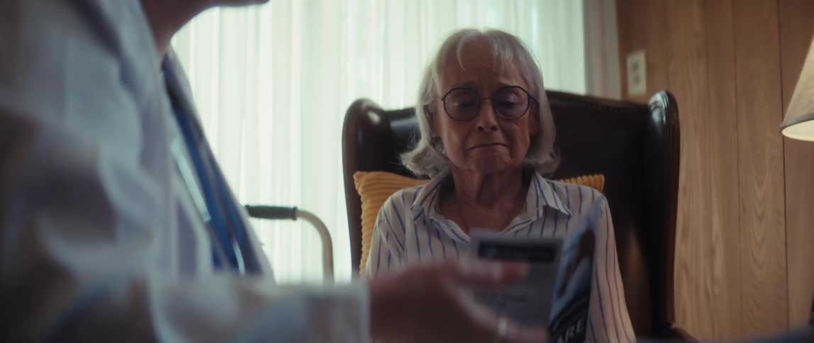 A somber elderly woman becomes tearful while reading a healthcare brochure presented by a doctor during an in-home visit
