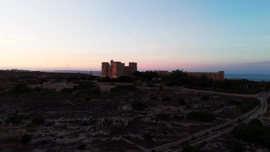 Aerial drone footage flies toward a historic stone fortress overlooking the sea, illuminated by the warm light of a beautiful sunset