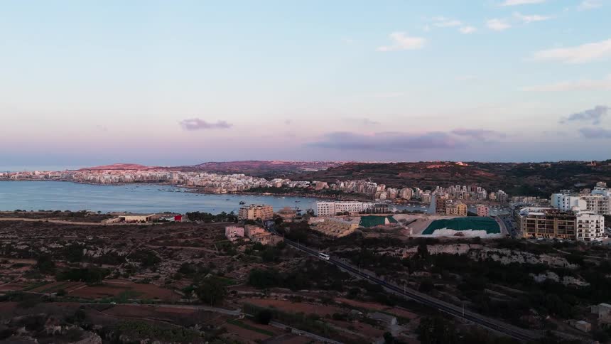An aerial view slowly pushes in over the countryside towards the coastal city of St. Paul's Bay, Malta, at sunset