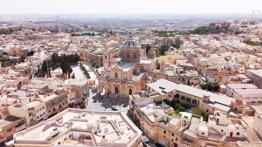 A stunning aerial view pans across a historic Maltese city, showcasing an ornate baroque-style church at its center