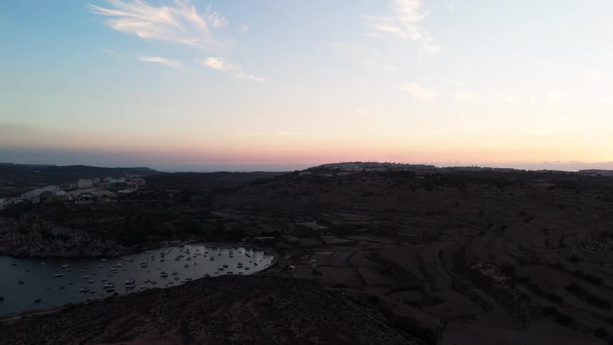 A tranquil aerial view shows a beautiful Mediterranean bay filled with boats and surrounded by rolling hills at sunset