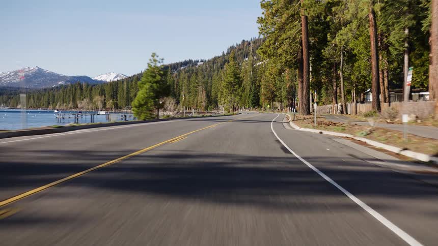 A scenic point-of-view drive on a lakeside road through a pine forest with snow-capped mountains in the distance