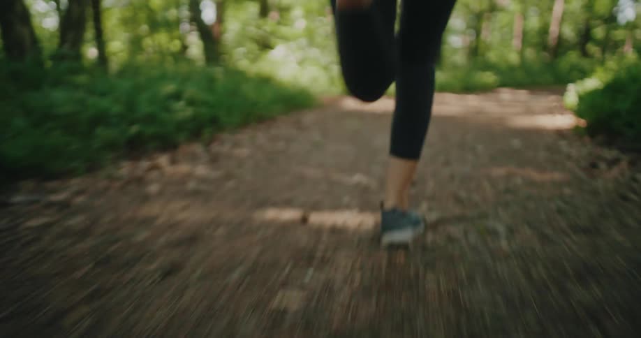 A low-angle follow shot captures the feet and legs of a person running along a dirt trail in a sunlit forest