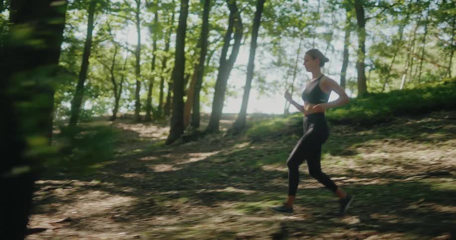 Side-view tracking shot of a fit woman running uphill on a sun-dappled forest trail