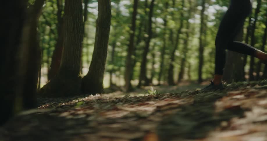 Side view slow motion close up of a woman's legs running on a forest trail with dappled sunlight