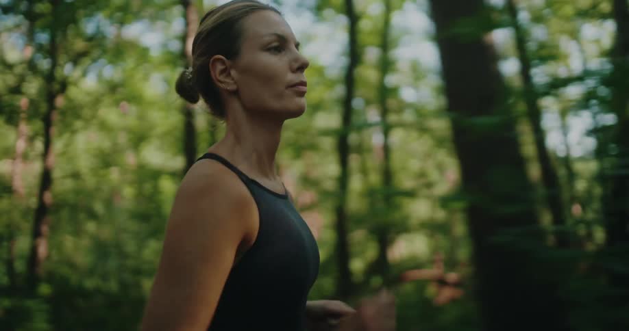 A fit woman runs through a lush green forest in the morning sunlight, with the camera following her from the side