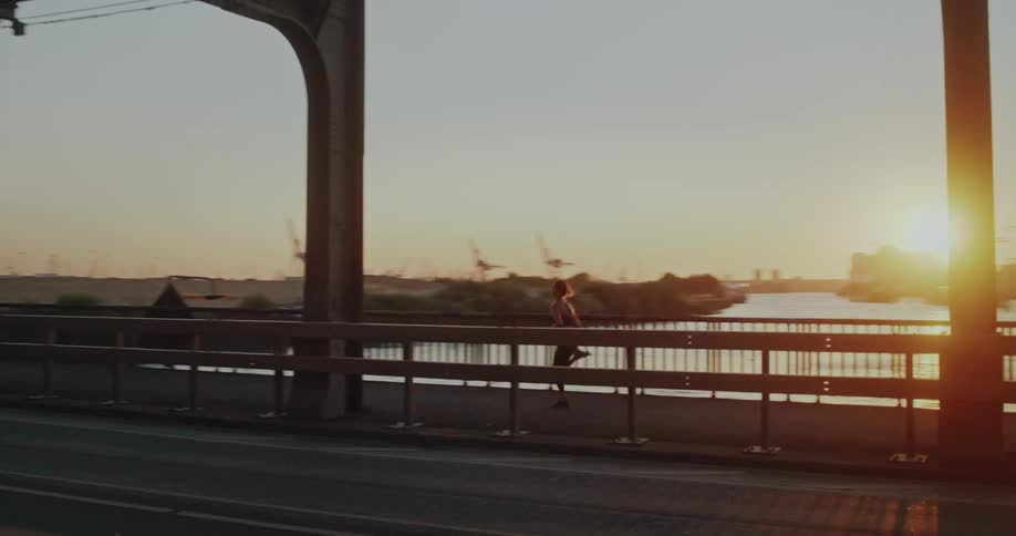 Tracking shot of an athletic woman running across a city bridge with an industrial backdrop at golden hour