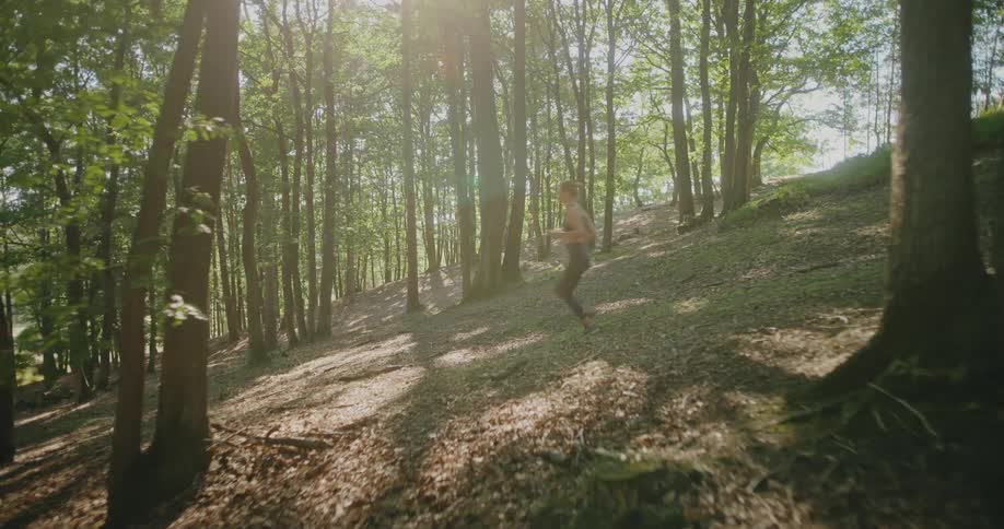 A fit young woman in sportswear runs uphill through a sun-drenched forest in slow motion