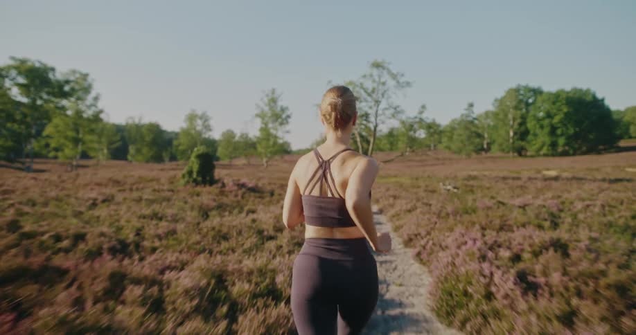 Slow motion shot from behind of a woman jogging on a trail in a purple heather field