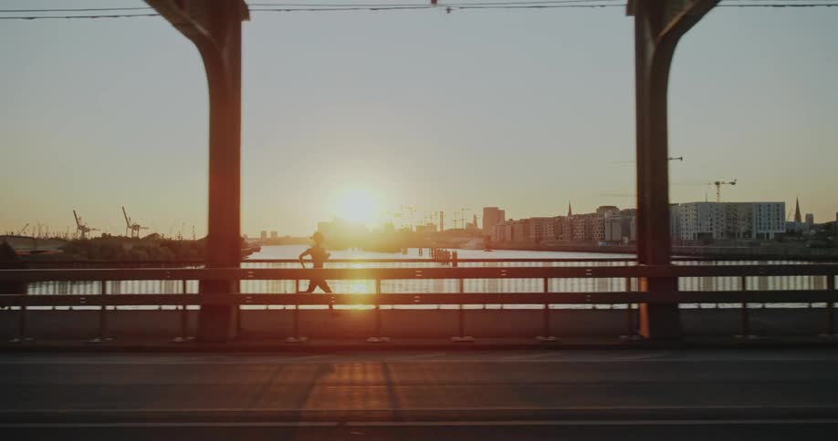 A woman jogs in slow motion across a city bridge with a beautiful sunrise reflecting on the water below